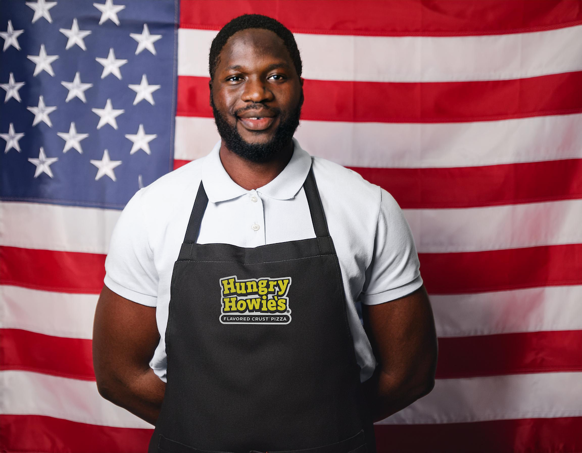African American veteran standing in front of a United States flag while smiling and wearing a Hungry Howie’s apron African American veteran standing in front of a United States flag while smiling and wearing a Hungry Howie's apron