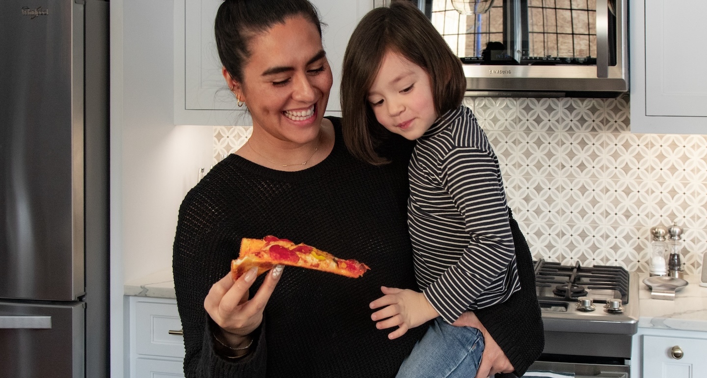 HH Lifestyle 1 Young woman holding her child, feeding her a slice of pizza in their kitchen