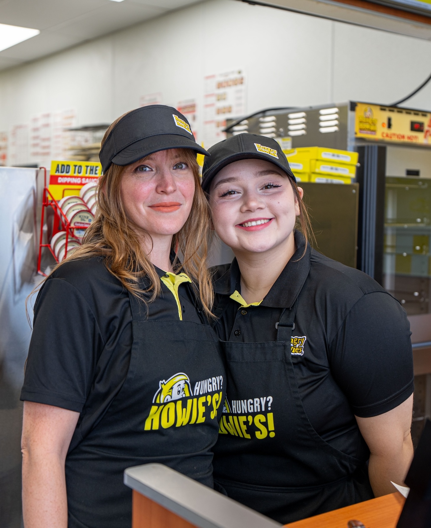 Two Hungry Howie’s employees standing at the register smiling at the camera Two Hungry Howie's employees standing at the register smiling at the camera