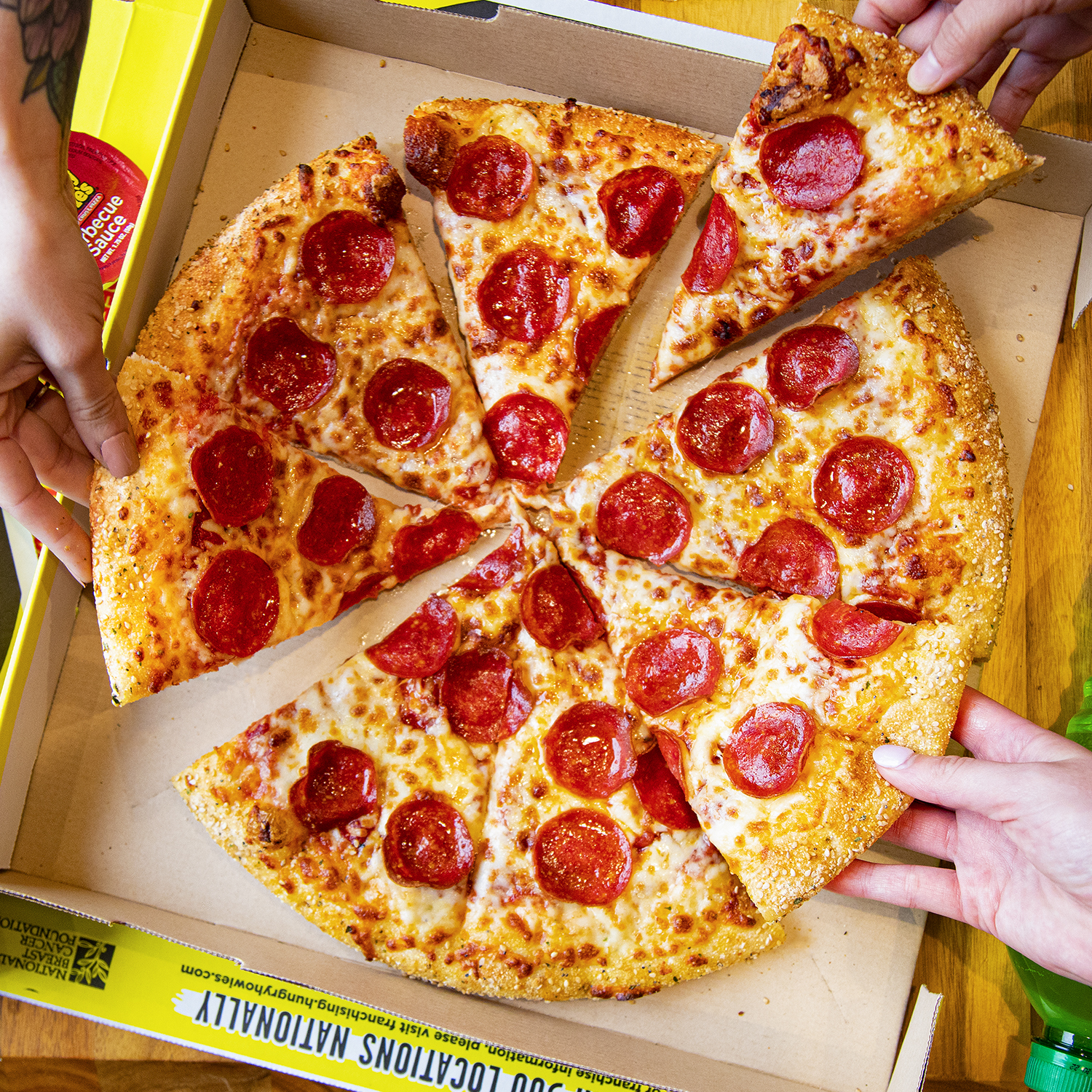 Overhead image of three people's hands pulling a large pepperoni pizza apart